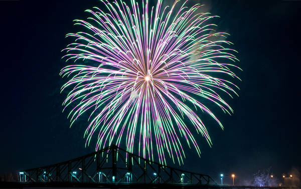 Un immense feu d'artifice multicolore éclate dans le ciel nocturne au-dessus d'un pont illuminé, créant une scène spectaculaire et vibrante.