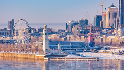 View of Montreal in winter from the Old Port with the Clock Tower in the foreground, then the ferris wheel behind it, and the snowy Montreal skyline in the background.