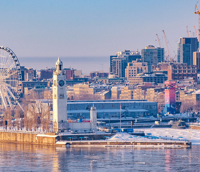 View of Montreal in winter from the Old Port with the Clock Tower in the foreground, then the ferris wheel behind it, and the snowy Montreal skyline in the background.