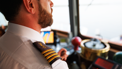 A ship captain in uniform, seen from behind, overlooking the navigation equipment and the St. Lawrence River from the deck of a ship.