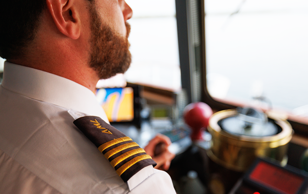 A ship captain in uniform, seen from behind, overlooking the navigation equipment and the St. Lawrence River from the deck of a ship.