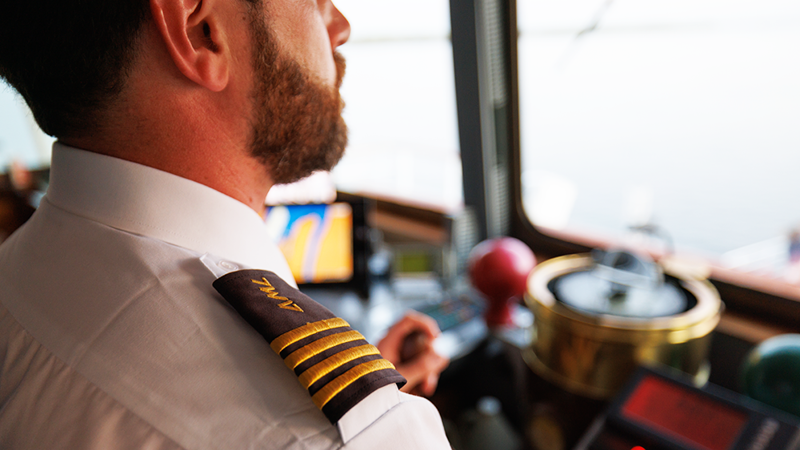 Un capitaine de navire en uniforme, vu de dos, surplombe l'équipement de navigation et le fleuve Saint-Laurent depuis le pont d'un navire.