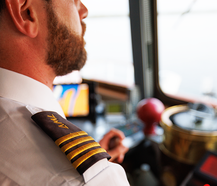 A ship captain in uniform, seen from behind, overlooking the navigation equipment and the St. Lawrence River from the deck of a ship.