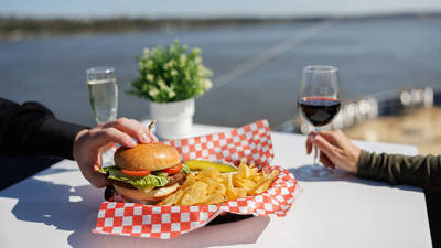 Une assiette contenant un hamburger, des chips et un cornichon sur une table blanche avec un motif à carreaux rouges et blancs. Une personne tient un verre de vin rouge, une autre touche le hamburger. En arrière-plan, une rivière et une plante en pot.