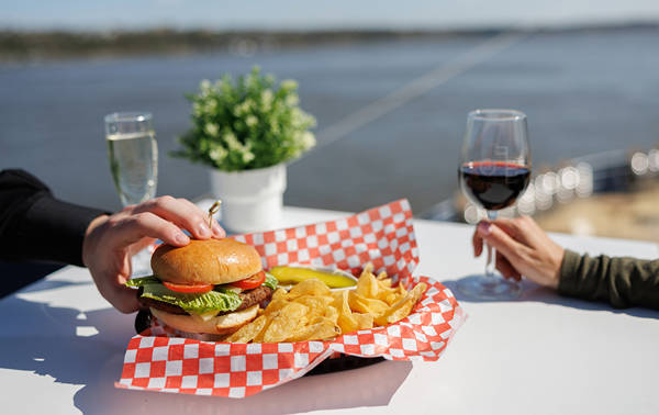 Une assiette contenant un hamburger, des chips et un cornichon sur une table blanche avec un motif à carreaux rouges et blancs. Une personne tient un verre de vin rouge, une autre touche le hamburger. En arrière-plan, une rivière et une plante en pot.