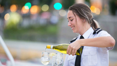 A smiling waitress pouring white wine into two glasses on a table.