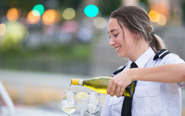 A smiling waitress pouring white wine into two glasses on a table.