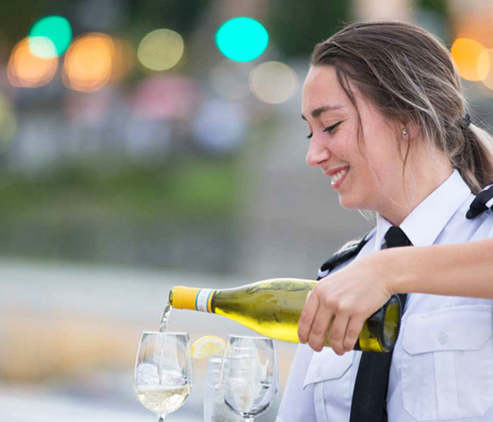 A smiling waitress pouring white wine into two glasses on a table.