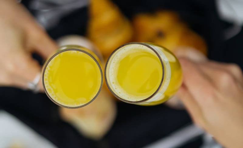 Two hands clink glasses filled with orange juice over a blurred background featuring plates and cutlery. The setting suggests a breakfast or brunch gathering.