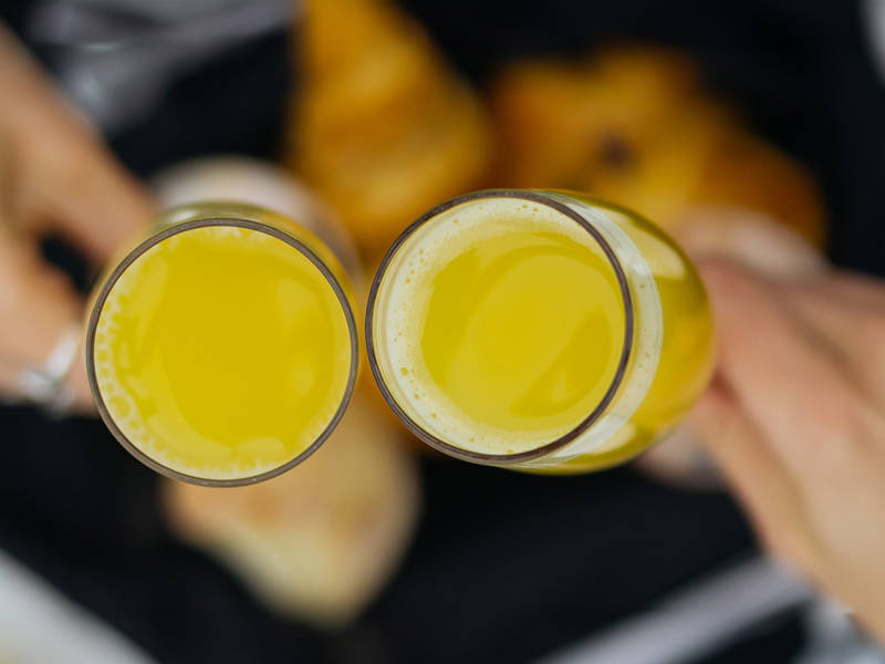 Two hands clink glasses filled with orange juice over a blurred background featuring plates and cutlery. The setting suggests a breakfast or brunch gathering.