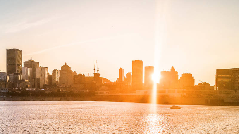 Une silhouette urbaine baignée dans une lumière dorée au coucher du soleil, avec des gratte-ciels en arrière-plan et une rivière scintillante au premier plan.