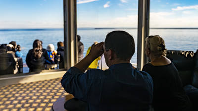 Un groupe de personnes se détend sur un bateau, admirant un vaste océan calme. Un homme au premier plan utilise des jumelles, tandis qu'une femme est assise à côté de lui. La scène est ensoleillée et paisible, avec un ciel bleu clair et des horizons lointains.