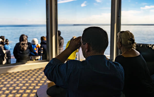 Un groupe de personnes se détend sur un bateau, admirant un vaste océan calme. Un homme au premier plan utilise des jumelles, tandis qu'une femme est assise à côté de lui. La scène est ensoleillée et paisible, avec un ciel bleu clair et des horizons lointains.