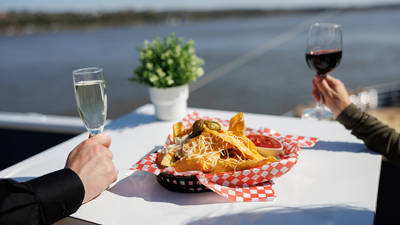 Une table blanche avec un panier de nachos garnis de fromage et de jalapeños, accompagnés de sauce rouge. Une personne tient un verre de vin rouge et une autre un verre de vin blanc, avec une rivière et une plante en arrière-plan.