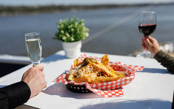 Une table blanche avec un panier de nachos garnis de fromage et de jalapeños, accompagnés de sauce rouge. Une personne tient un verre de vin rouge et une autre un verre de vin blanc, avec une rivière et une plante en arrière-plan.