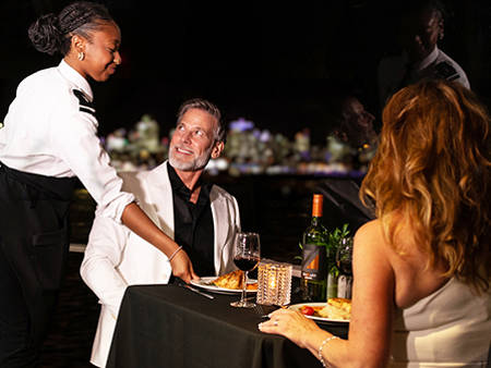 Un couple élégamment habillé dîne à une table romantique, servi par une serveuse souriante. La table est décorée avec des bougies, du vin et des assiettes de plats raffinés, avec une vue nocturne floue en arrière-plan.