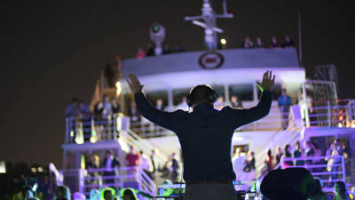 Un DJ anime une soirée festive sur le pont d'un bateau, les bras levés, avec une foule de participants et des lumières colorées éclairant la scène pendant la nuit.