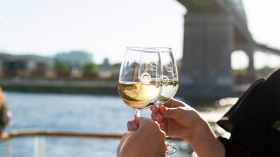 Deux personnes trinquent avec des verres de vin blanc sur une terrasse extérieure, avec une rivière, un pont et des bâtiments en arrière-plan par une journée ensoleillée.