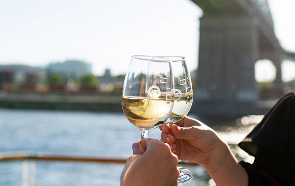 Deux personnes trinquent avec des verres de vin blanc sur une terrasse extérieure, avec une rivière, un pont et des bâtiments en arrière-plan par une journée ensoleillée.