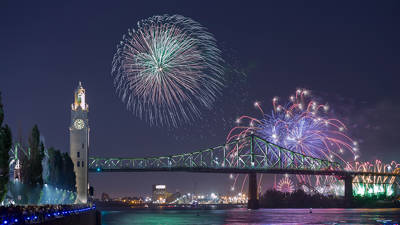 A spectacular fireworks display lights up the night sky above the Jacques-Cartier Bridge in Montreal. The Clock Tower stands on the left, while vibrant colors reflect on the Saint Lawrence River. A crowd watches the show from the dock.