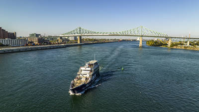 Un bateau de croisière navigue sur une rivière sous un grand pont vert, avec des bâtiments urbains et un ciel clair en arrière-plan.