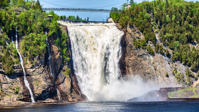 Montmorency Falls in the summer cascading into the Montmorency River below, surrounded by trees with a suspended footbridge above