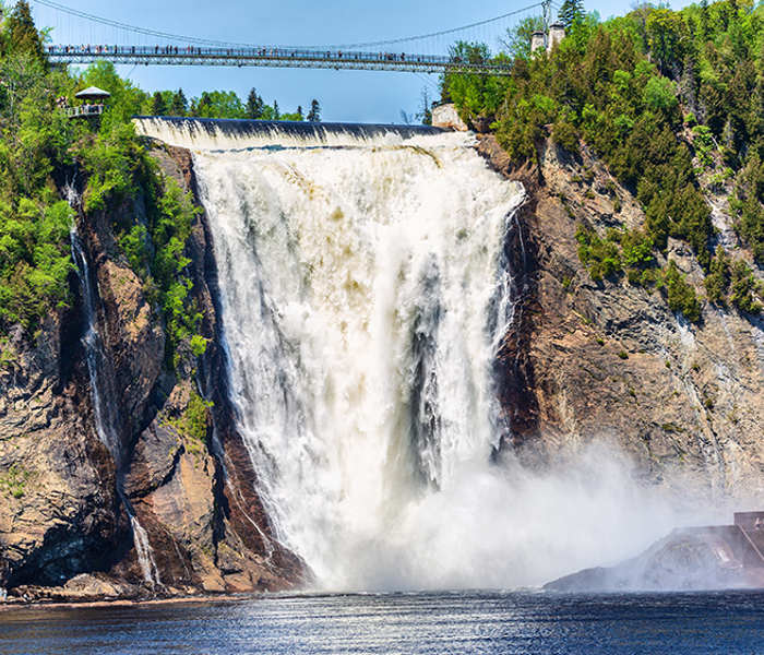 Montmorency Falls in the summer cascading into the Montmorency River below, surrounded by trees with a suspended footbridge above