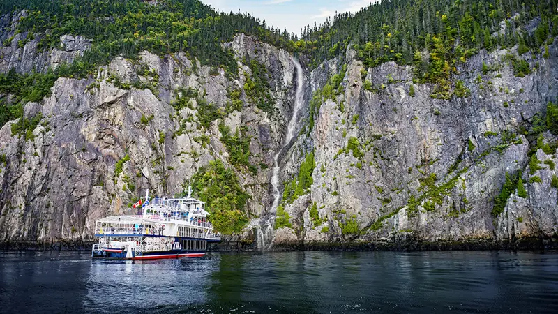 A large tour boat sails near towering rocky cliffs with lush green trees, in front of a cascading waterfall that flows into the calm water below. The scene is tranquil, featuring clear skies and reflections on the water's surface.