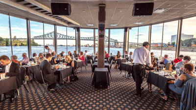 Salle principale du bateau avec de grandes fenêtres où des clients soupent tout en admirant la vue sur le fleuve Saint-Laurent