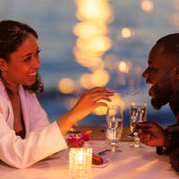 Un couple partage un moment romantique lors d'un dîner aux chandelles près de l'eau. La femme, souriante, donne une bouchée de dessert à l'homme, avec des lumières floues scintillant en arrière-plan.