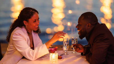 Un couple partage un moment romantique lors d'un dîner aux chandelles près de l'eau. La femme, souriante, donne une bouchée de dessert à l'homme, avec des lumières floues scintillant en arrière-plan.