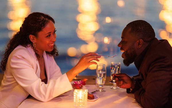 Un couple partage un moment romantique lors d'un dîner aux chandelles près de l'eau. La femme, souriante, donne une bouchée de dessert à l'homme, avec des lumières floues scintillant en arrière-plan.