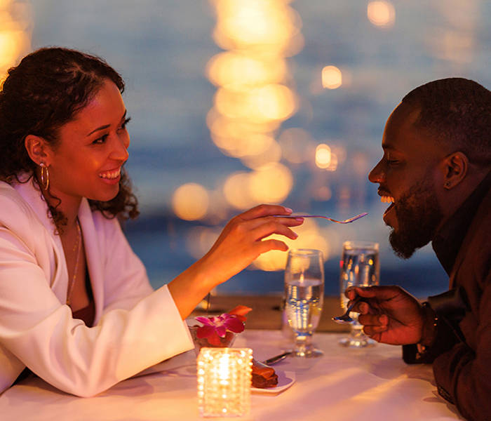 Un couple partage un moment romantique lors d'un dîner aux chandelles près de l'eau. La femme, souriante, donne une bouchée de dessert à l'homme, avec des lumières floues scintillant en arrière-plan.