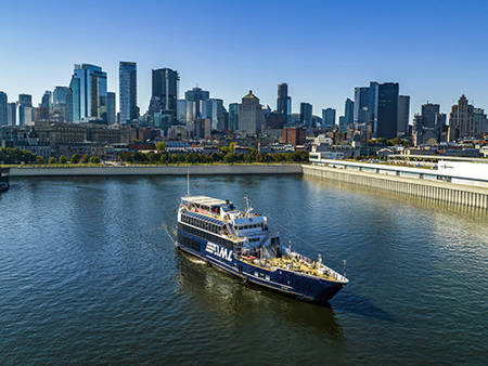 A ship with "AML" written on its side sails on a calm river. The background features a city skyline with modern skyscrapers under a clear blue sky.