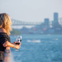 Une femme blonde, de dos, tenant un verre de vin rouge, regardant une rivière avec un pont et des gratte-ciel en arrière-plan par une journée ensoleillée.