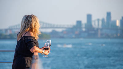 Une femme blonde, de dos, tenant un verre de vin rouge, regardant une rivière avec un pont et des gratte-ciel en arrière-plan par une journée ensoleillée.