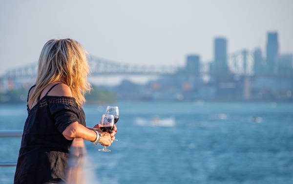 Une femme blonde, de dos, tenant un verre de vin rouge, regardant une rivière avec un pont et des gratte-ciel en arrière-plan par une journée ensoleillée.