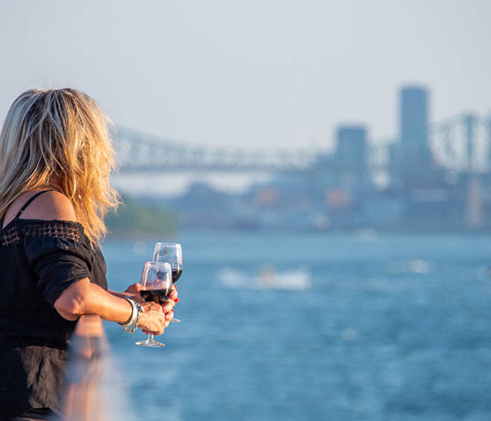 Une femme blonde, de dos, tenant un verre de vin rouge, regardant une rivière avec un pont et des gratte-ciel en arrière-plan par une journée ensoleillée.