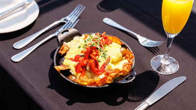 Cassolette gourmande comprise dans le brunch avec œufs brouillés, pommes de terre grelots, poivrons rôtis, fromage en grains, bacon, saucisses et sauce hollandaise accompagnée d'un verre de mimosa 