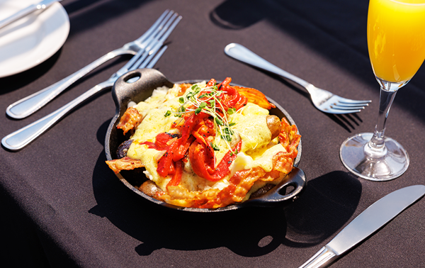 Cassolette gourmande comprise dans le brunch avec œufs brouillés, pommes de terre grelots, poivrons rôtis, fromage en grains, bacon, saucisses et sauce hollandaise accompagnée d'un verre de mimosa 