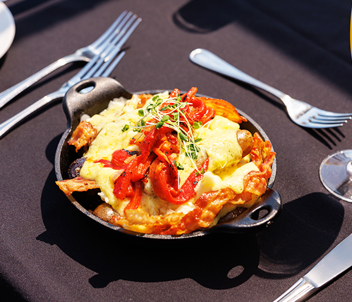 Cassolette gourmande comprise dans le brunch avec œufs brouillés, pommes de terre grelots, poivrons rôtis, fromage en grains, bacon, saucisses et sauce hollandaise accompagnée d'un verre de mimosa 