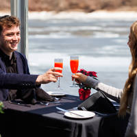 A couple toasts with red cocktails during an elegant meal near a window overlooking a river, with a decorated Christmas tree in the background.