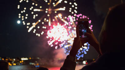  Une personne tient un smartphone pour capturer des feux d'artifice colorés illuminant le ciel nocturne, avec des reflets sur l'eau et une ville en arrière-plan.