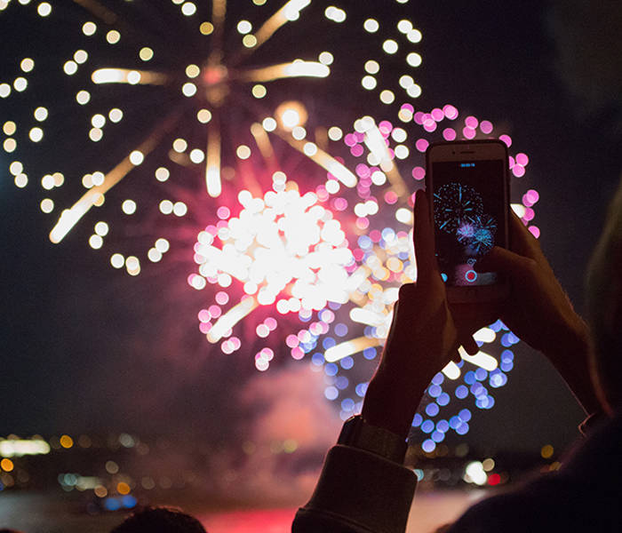  Une personne tient un smartphone pour capturer des feux d'artifice colorés illuminant le ciel nocturne, avec des reflets sur l'eau et une ville en arrière-plan.