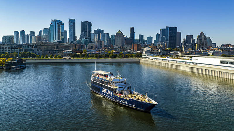 Un bateau de croisière naviguant sur une rivière, avec une vue panoramique sur une ville avec de hauts gratte-ciels en arrière-plan sous un ciel dégagé.