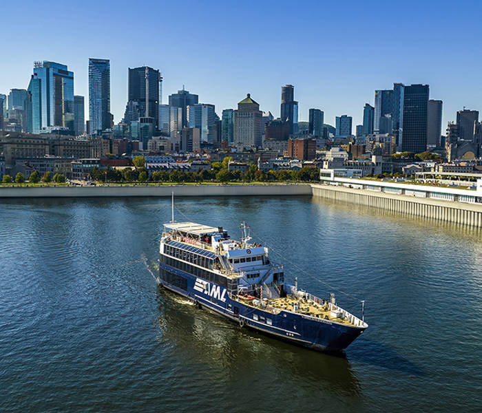 Un bateau de croisière naviguant sur une rivière, avec une vue panoramique sur une ville avec de hauts gratte-ciels en arrière-plan sous un ciel dégagé.