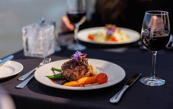 Un plat gastronomique composé d'un morceau de viande juteux, accompagné de légumes, d'une purée et d'une sauce, décoré avec des fleurs comestibles. Le repas est servi sur une table élégante avec un verre de vin rouge.