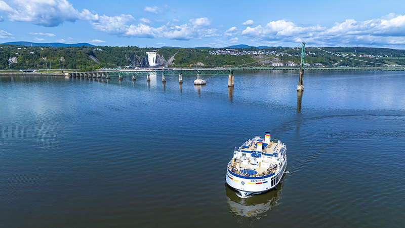 Aerial view of the AML Louis Jolliet sailing on the St. Lawrence River, with the Île d'Orléans bridge and Montmorency Falls in the background.