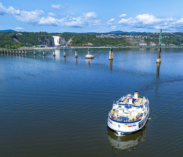 Aerial view of the AML Louis Jolliet sailing on the St. Lawrence River, with the Île d'Orléans bridge and Montmorency Falls in the background.