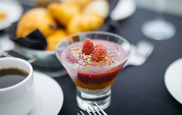 Un dessert élégant dans un verre, composé de couches de mousse ou de purée de fruits rouges, garni de framboises fraîches et de miettes de noix ou de biscuits, accompagné d'une tasse de café et de croissants en arrière-plan.